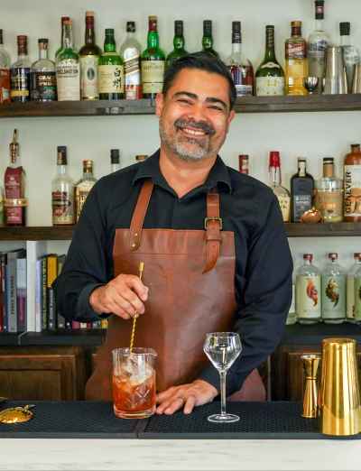 Smiling bartender stirring a cocktail behind a bar with shelves of liquor bottles.