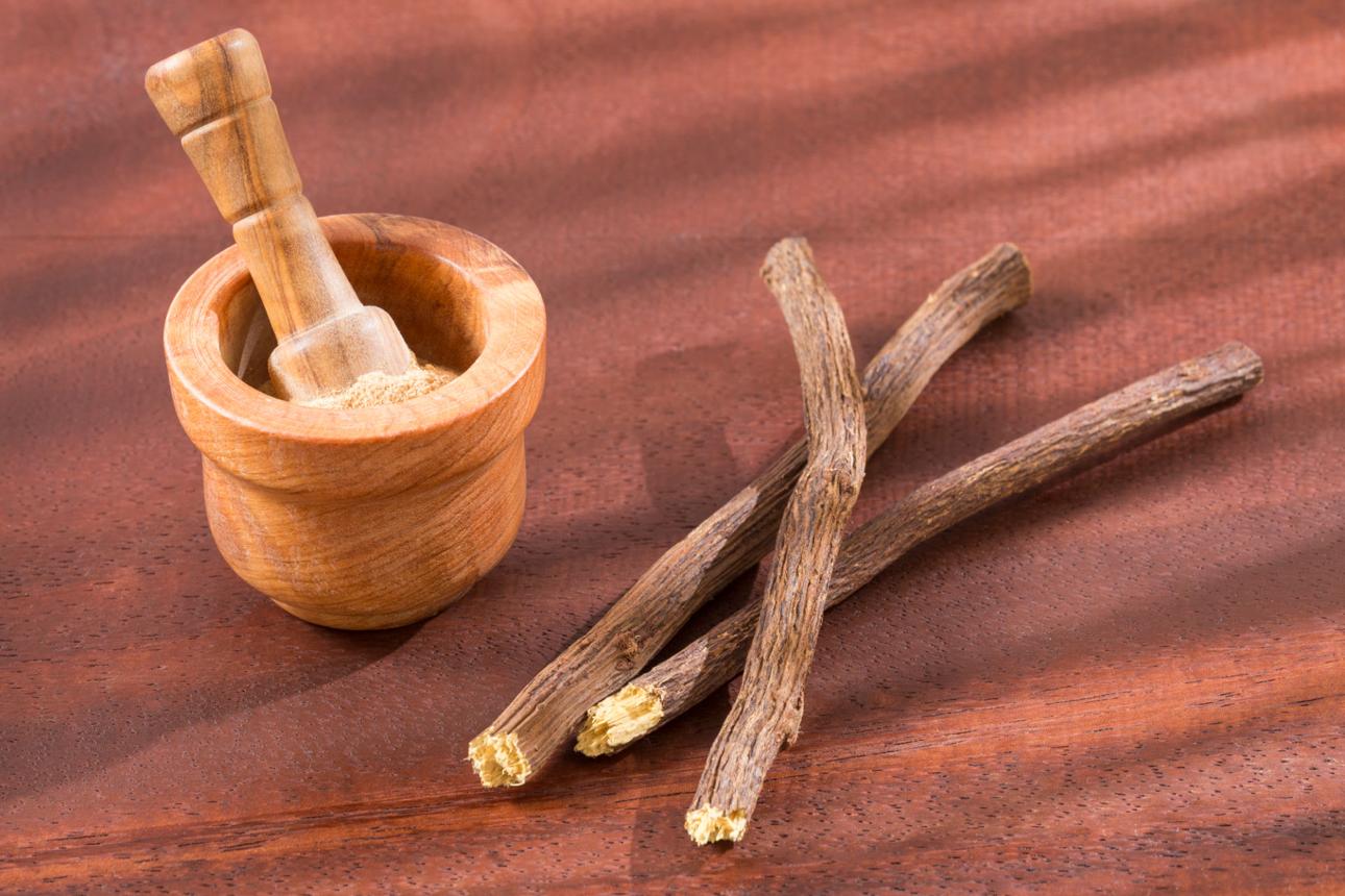 A wooden mortar and pestle with ground powder and several licorice root sticks on a dark wood surface.