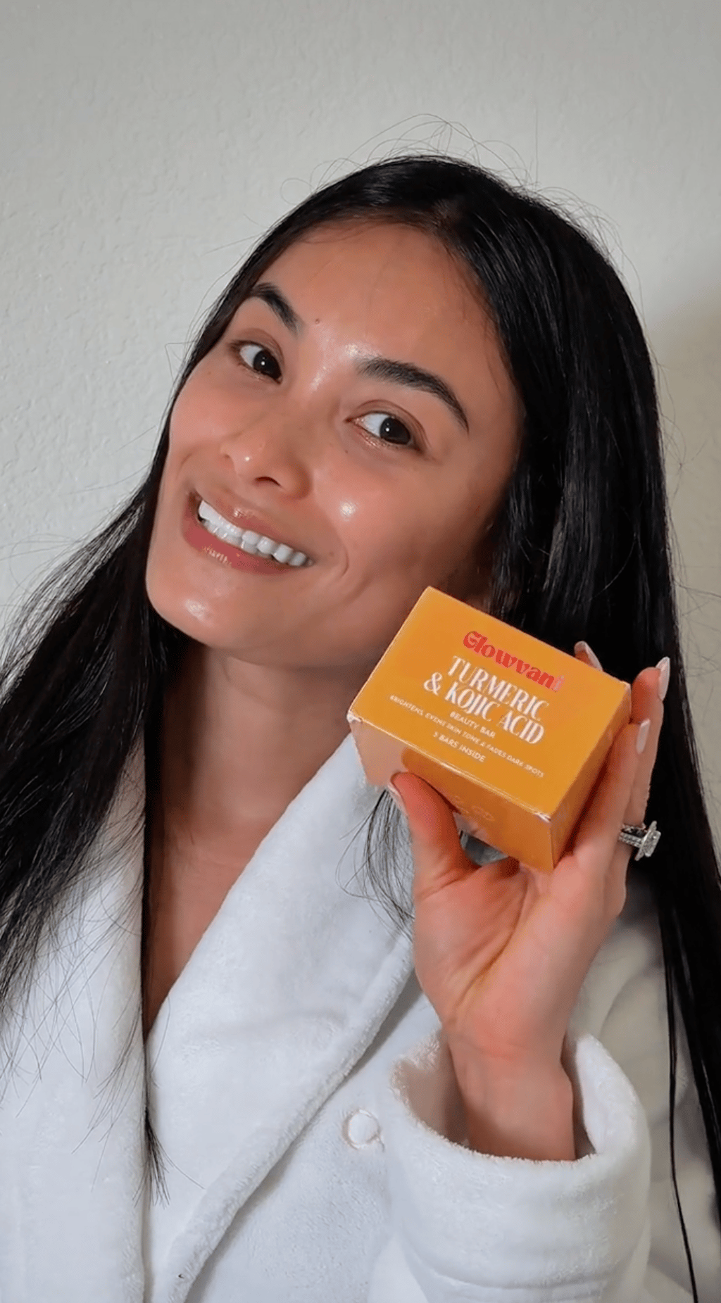 A smiling woman in a white robe holds a small orange box of Turmeric & Kojic Acid soap.