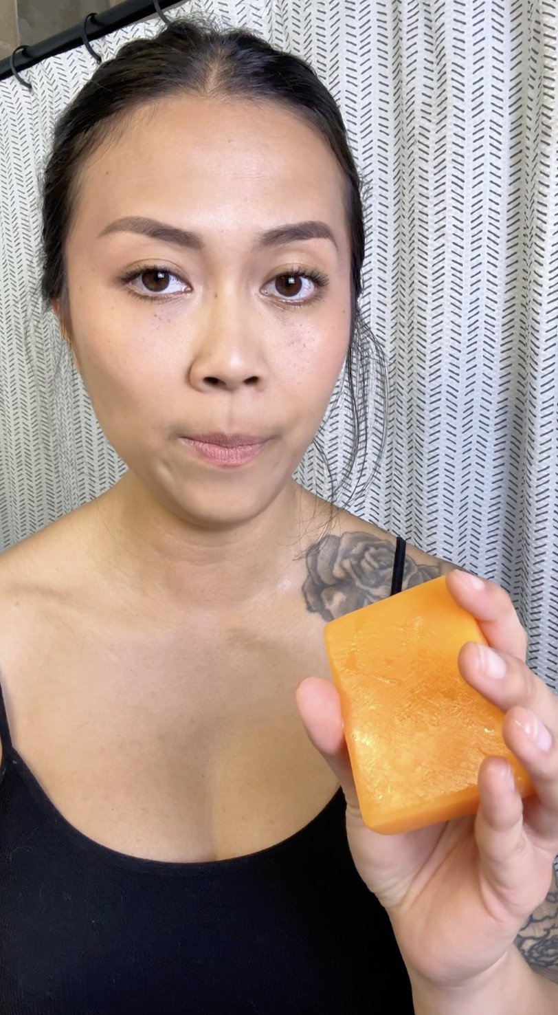 Person holding an orange soap bar in front of a patterned shower curtain.