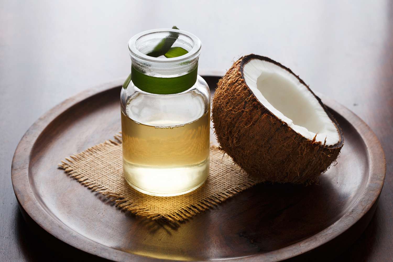 Glass jar of oil and half coconut on a wooden tray.