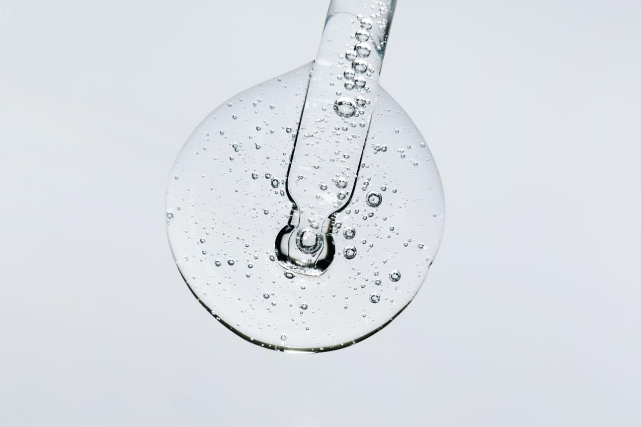 A close-up of a glass dropper in a circular drop of clear, bubbly serum against a white background.