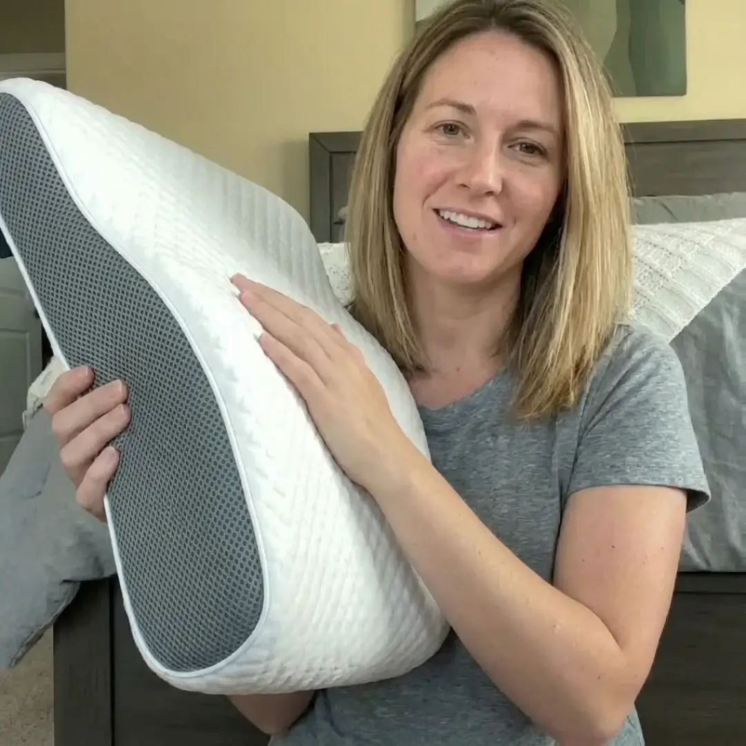 A smiling woman in a grey t-shirt holds up a white and grey contoured pillow.