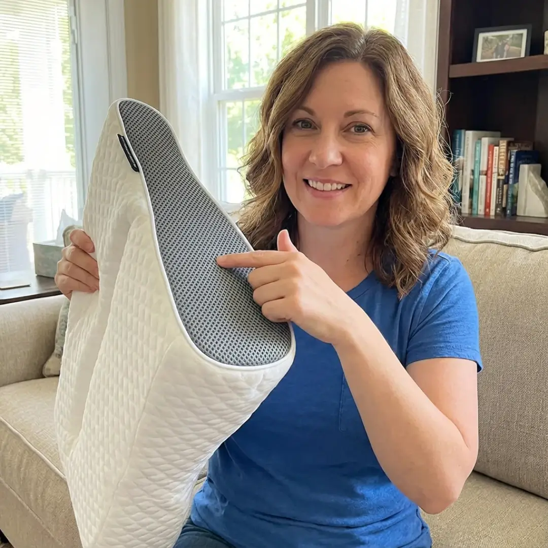 A smiling woman in a blue shirt holds up a white and grey pillow, pointing to it.
