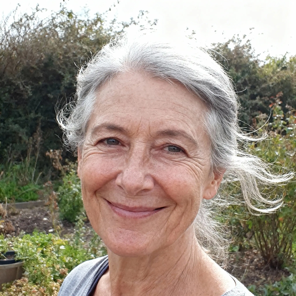 Smiling person with grey hair outdoors, surrounded by greenery.
