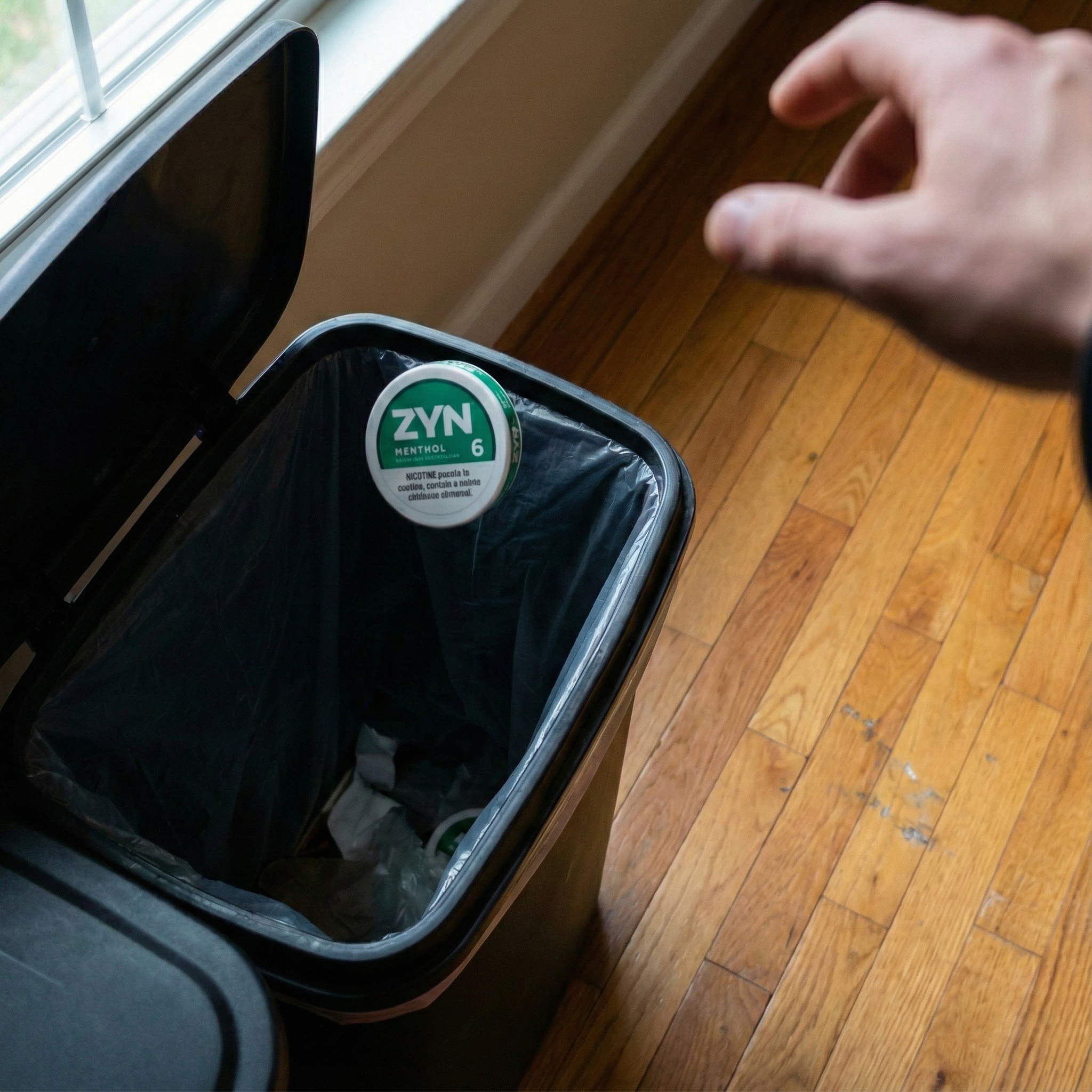 Hand throwing a ZYN can into a trash bin.