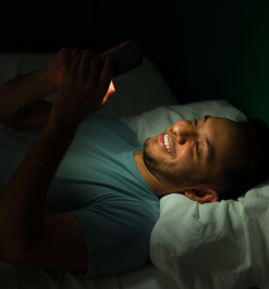 Man lying in bed, smiling at a smartphone in the dark.