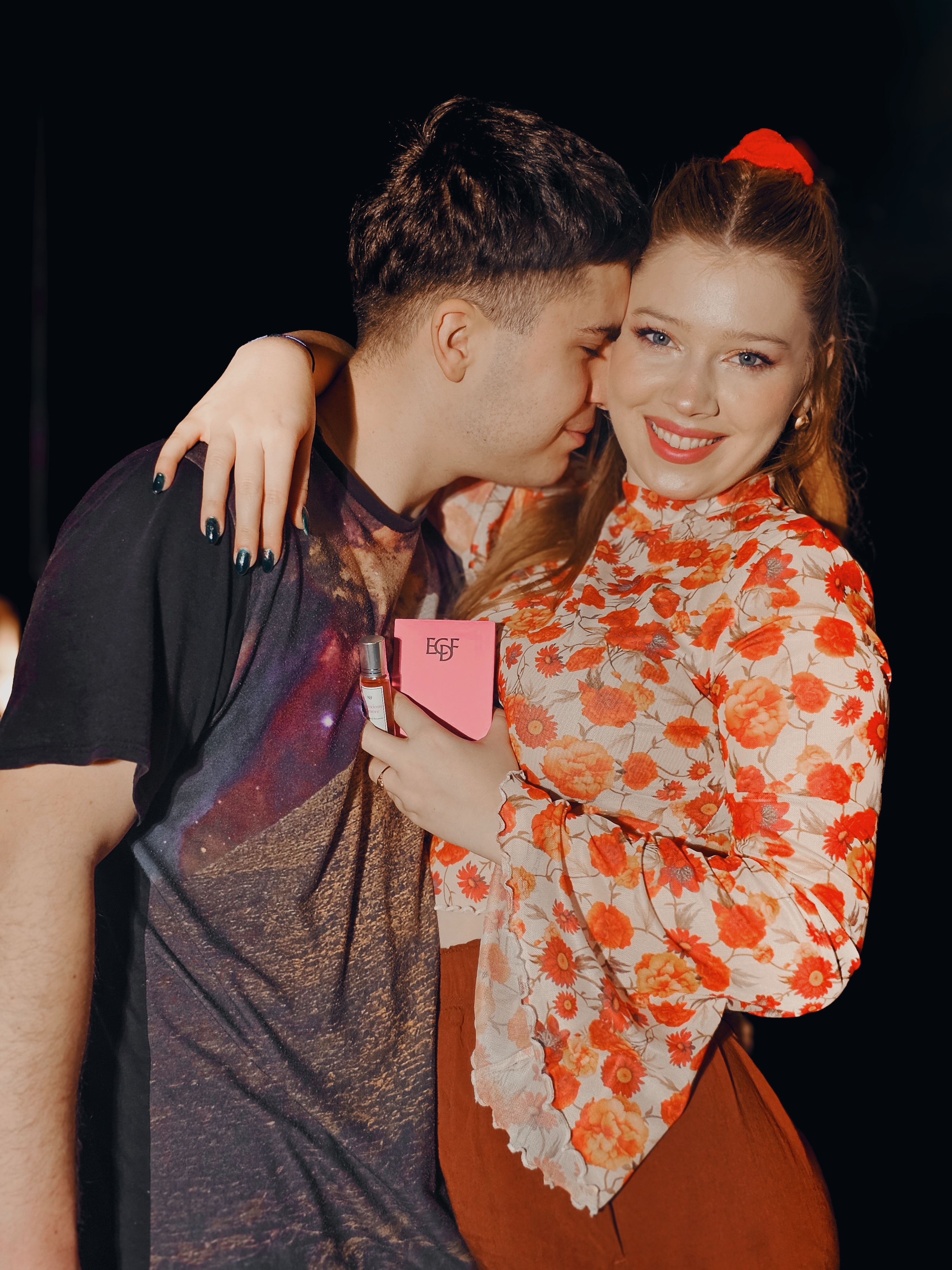 Smiling couple embracing, holding a pink box and small bottle.
