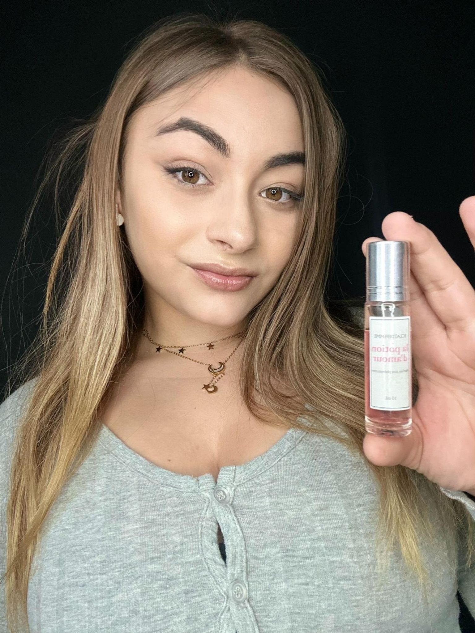 Person holding a small perfume bottle, wearing a grey top and star necklace.