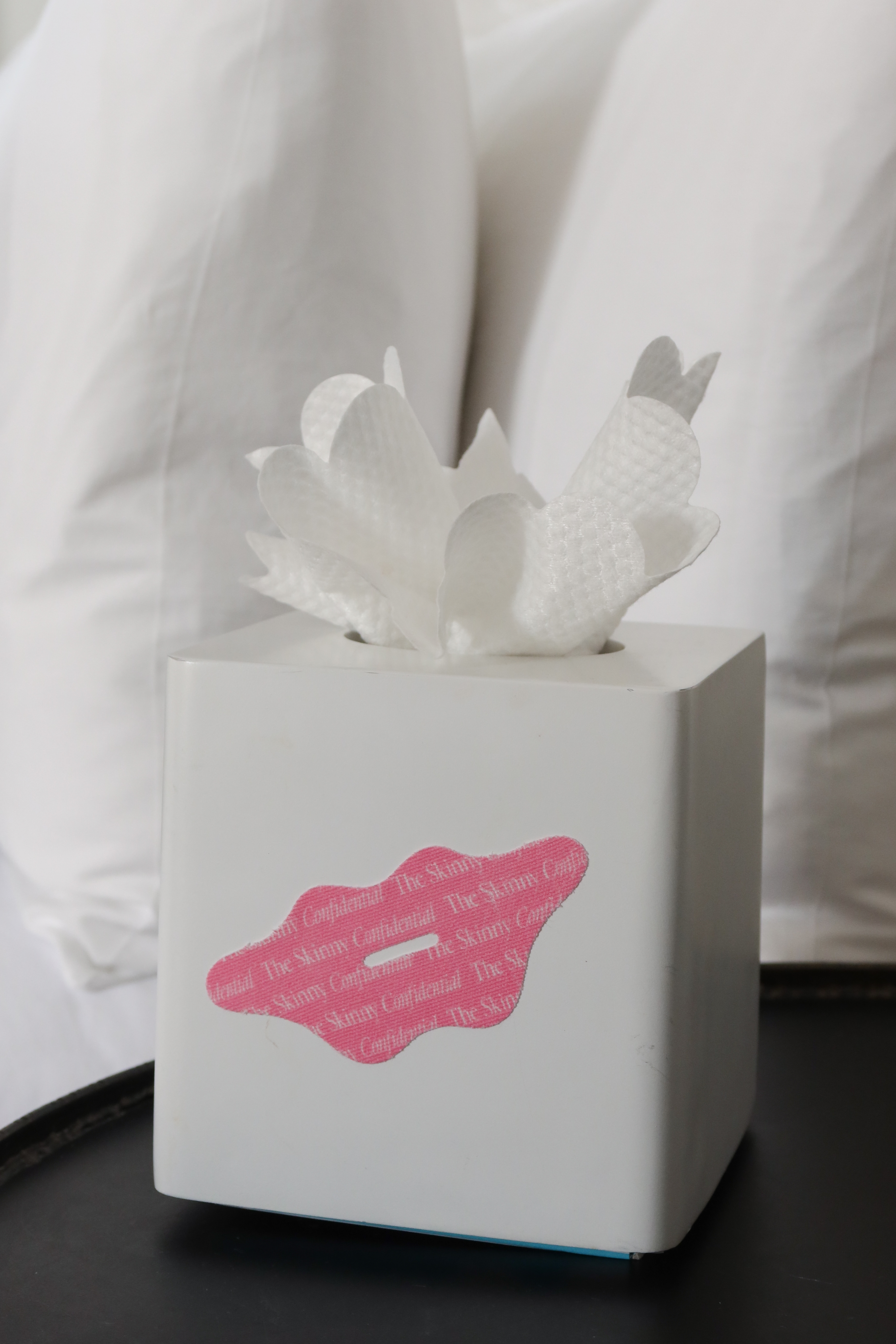 A white tissue box holder with a pink lip-shaped sticker sits on a dark table in front of pillows.