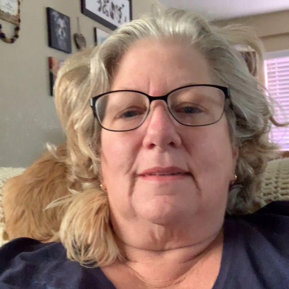 Person with glasses sitting indoors, a wall with frames behind.