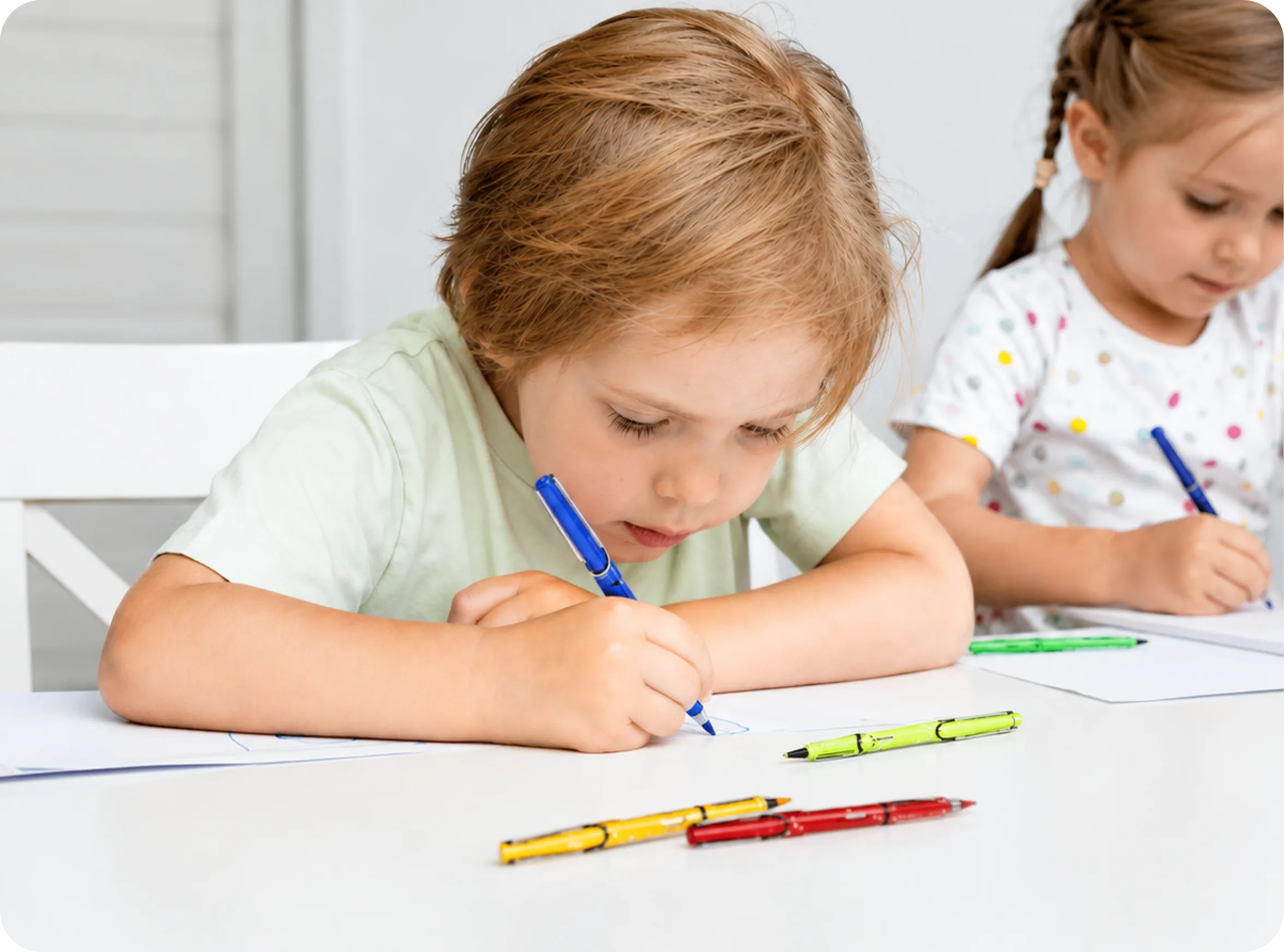 A young boy and girl sit at a white table, focused on drawing with colorful markers.