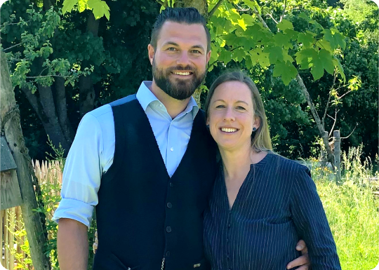 A man and woman smiling outdoors with trees in the background.