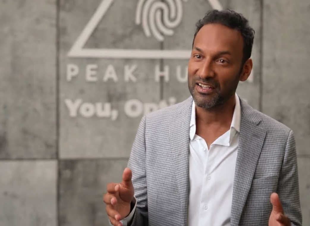 A person in a blazer speaking in front of a wall with Peak Humanity logo.