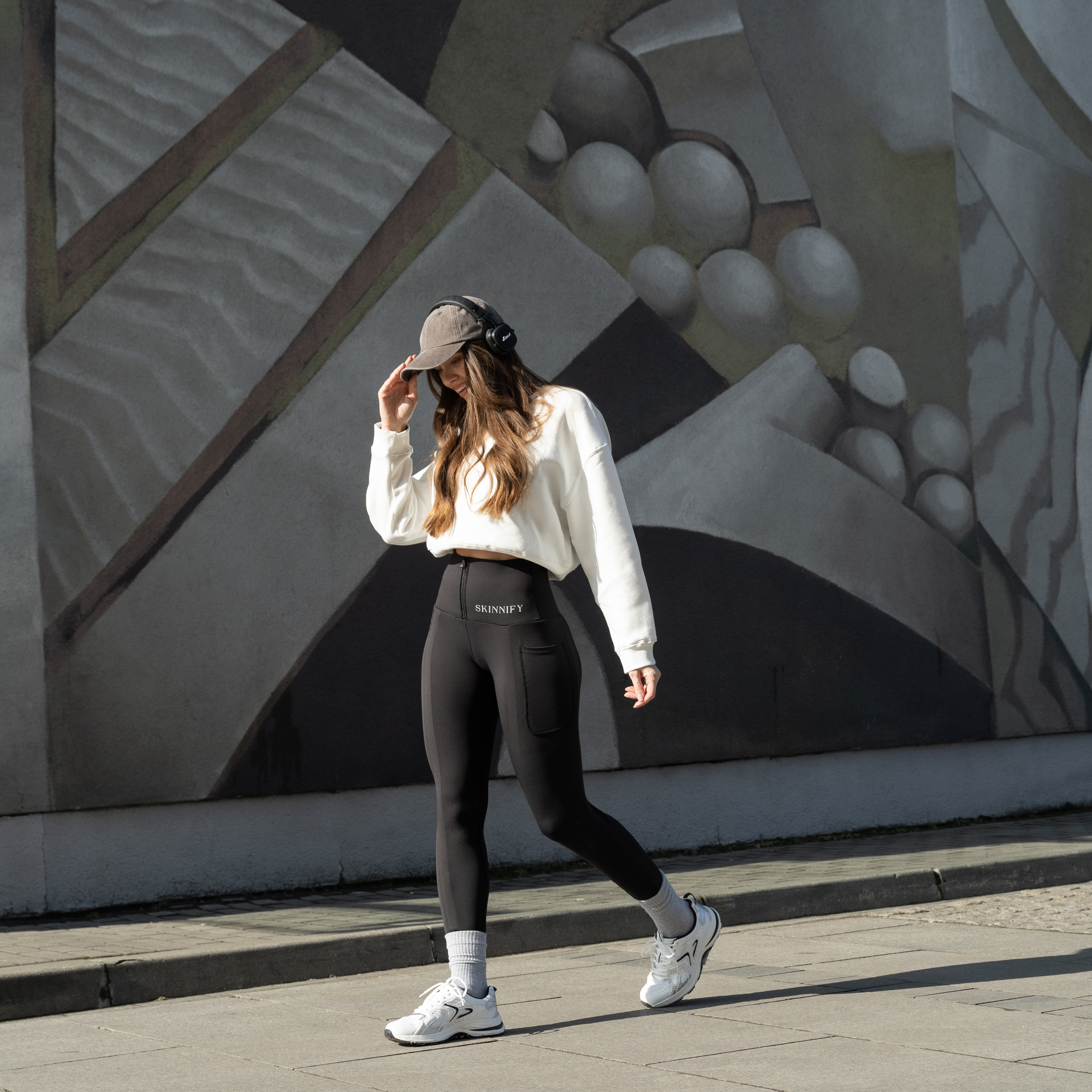 Woman in athletic wear walking past a mural, wearing headphones and a cap.