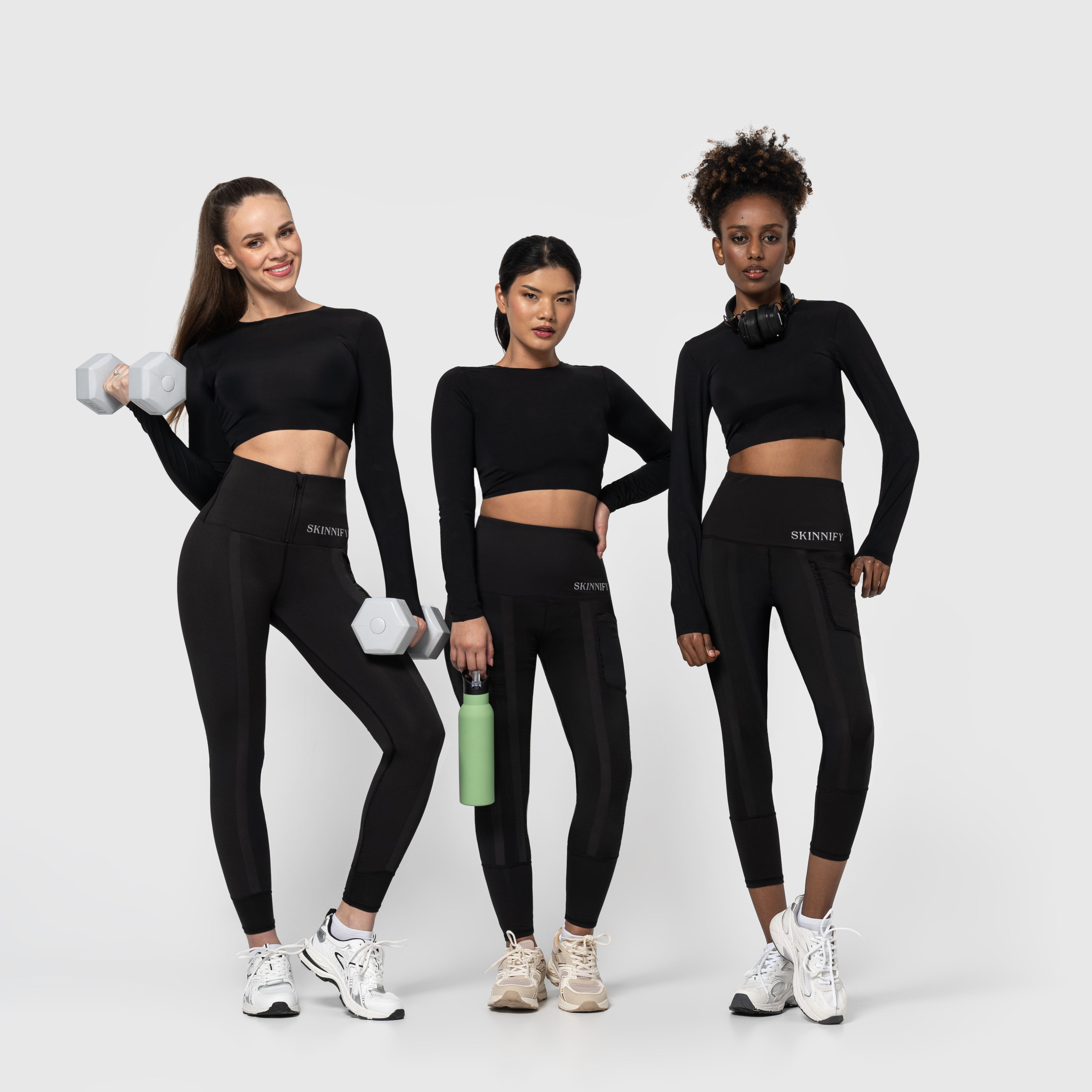 Three women in black activewear posing confidently with dumbbells and water bottle.