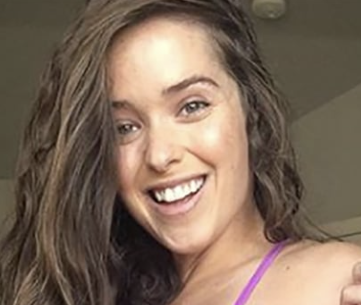 A woman smiles brightly, with long wavy hair, indoors.