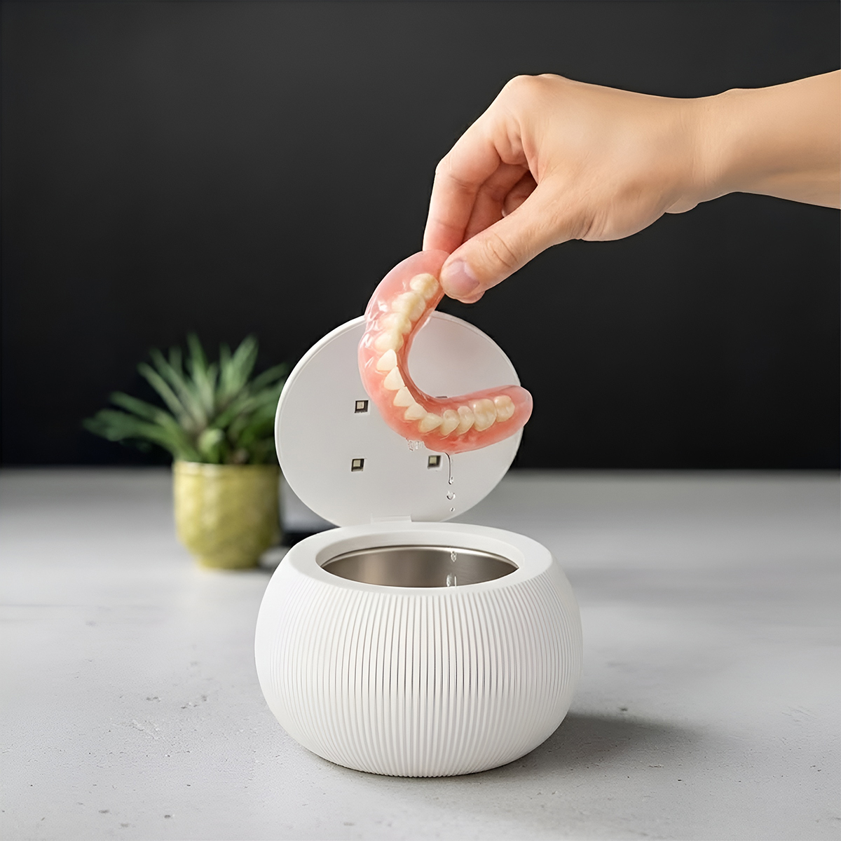 Hand holding dentures over a cleaning device on a table.