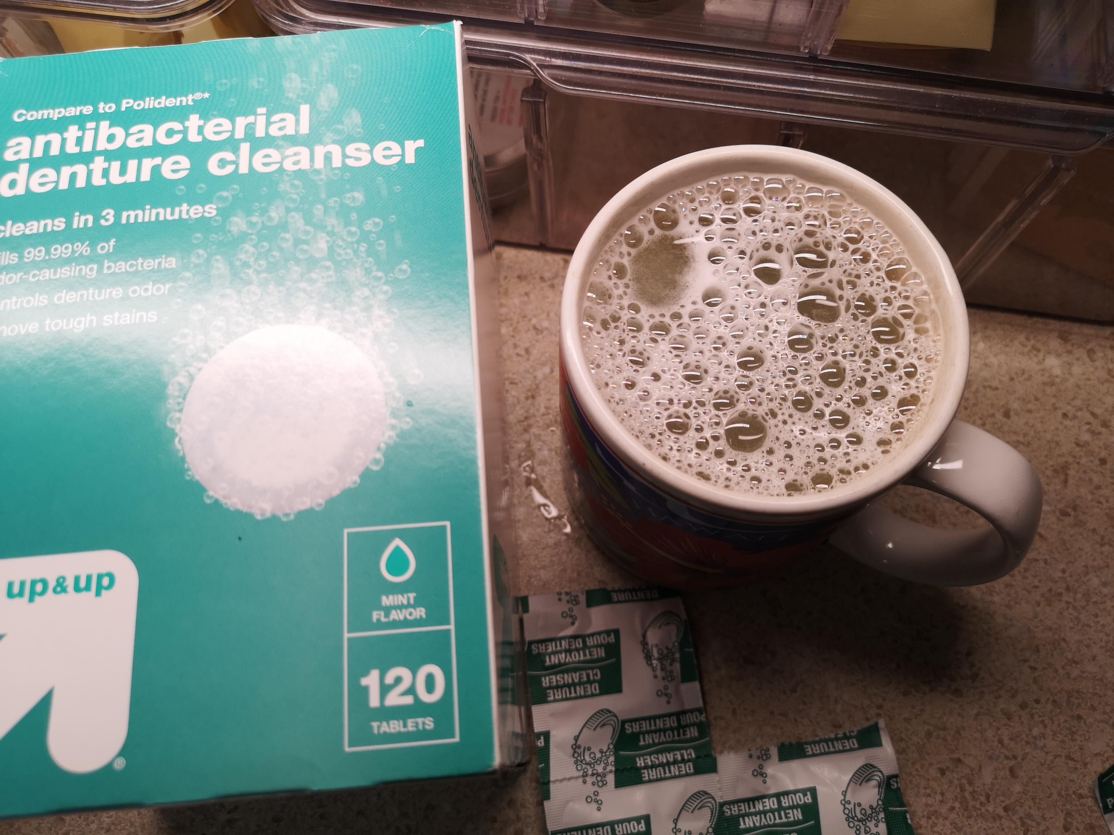 A box of denture cleanser tablets next to a mug with a fizzy, bubbling cleaning solution.