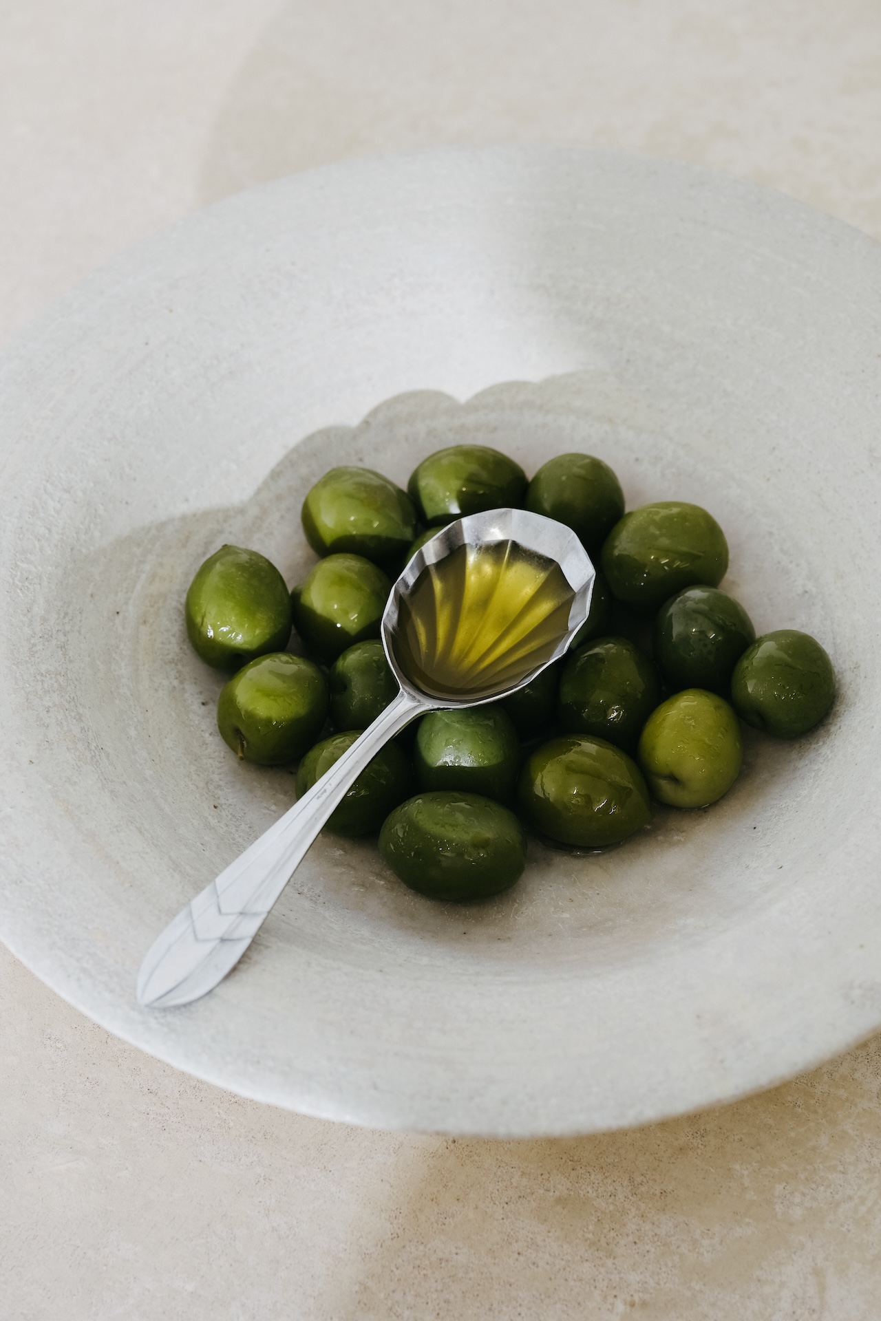 A spoonful of olive oil rests on a pile of green olives in a rustic, light-colored bowl.