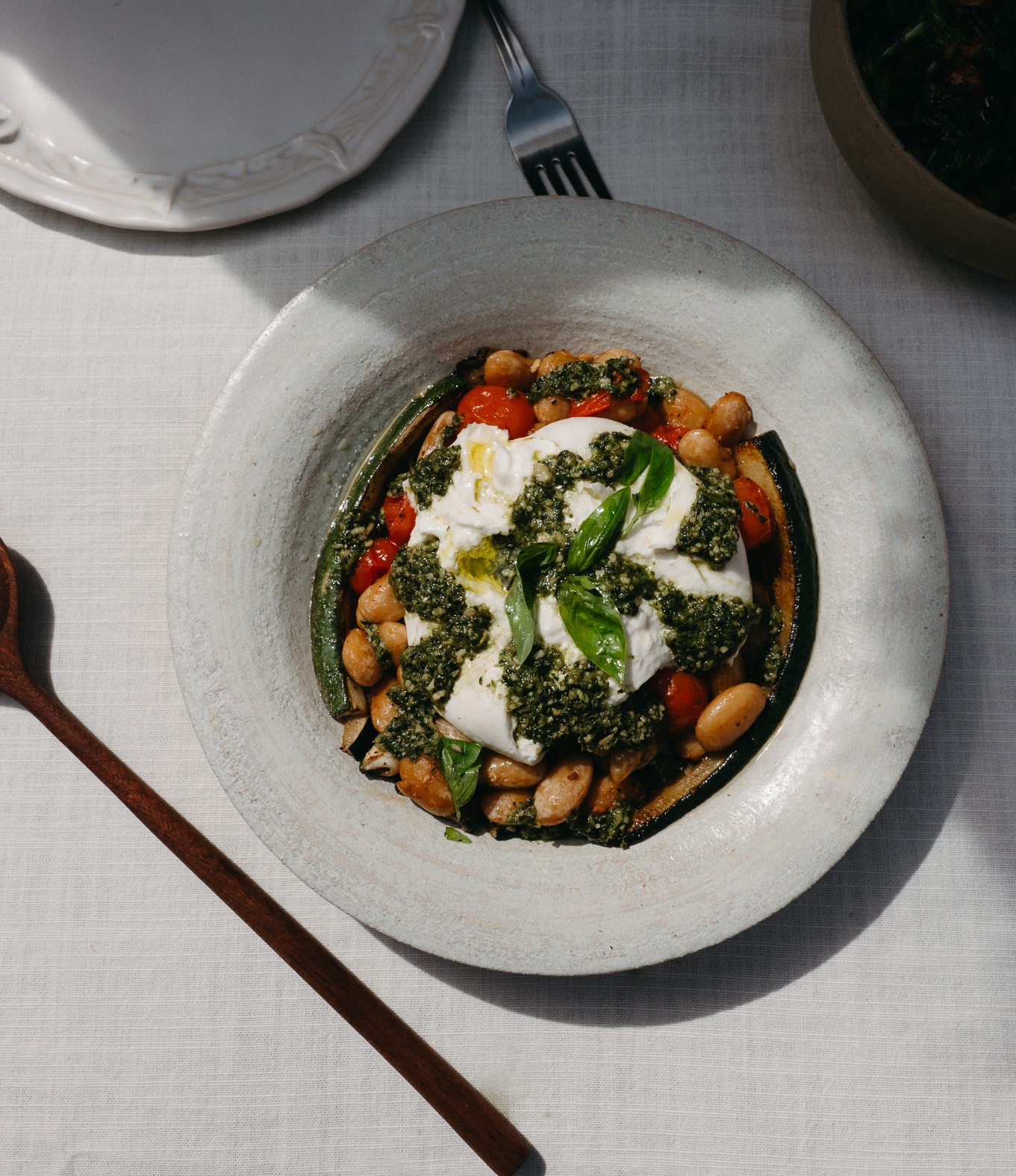 A rustic bowl of white beans, tomatoes, and burrata cheese, topped with pesto and fresh basil.