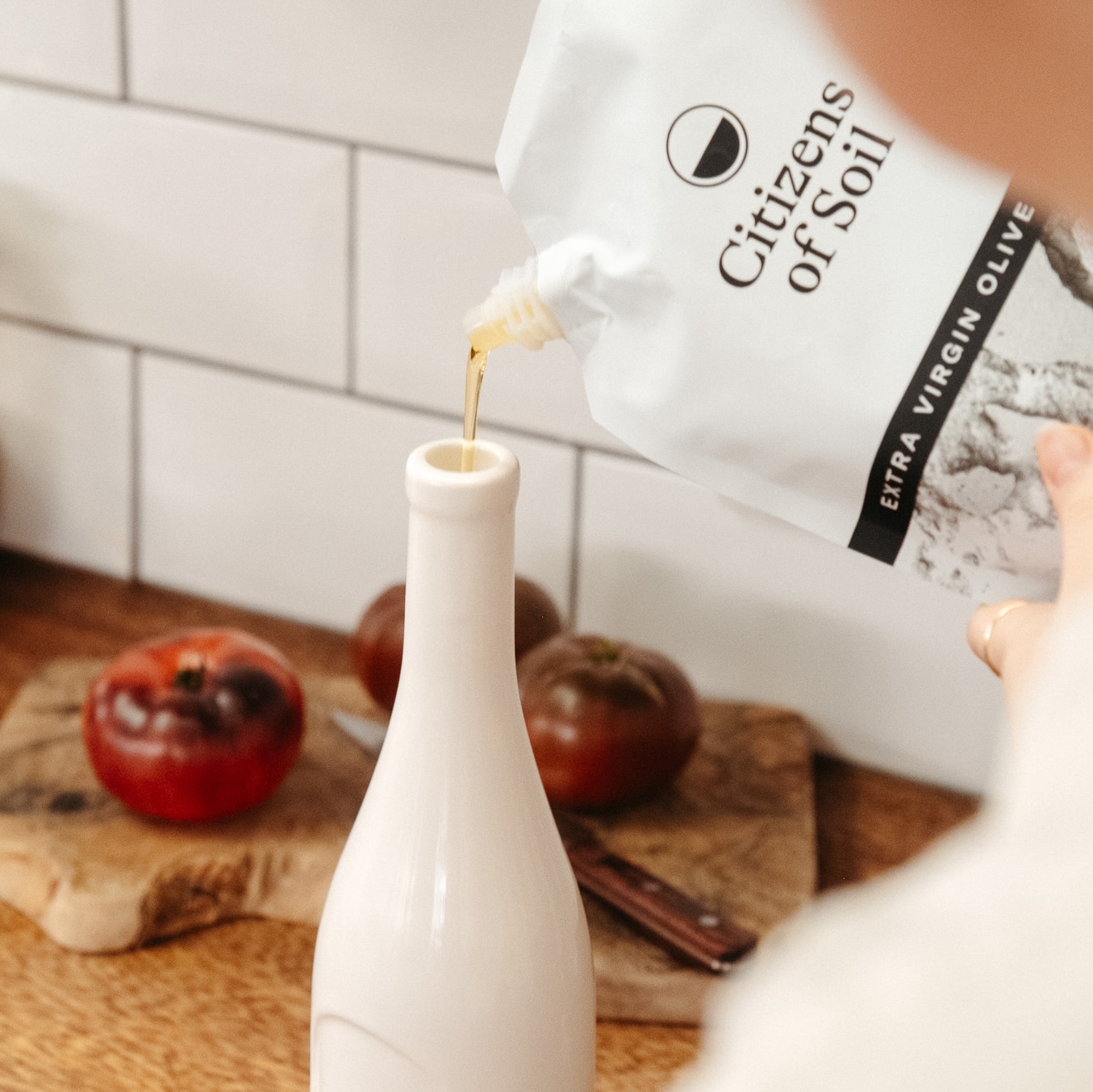 A person pours extra virgin olive oil from a Citizens of Soil pouch into a white ceramic bottle.