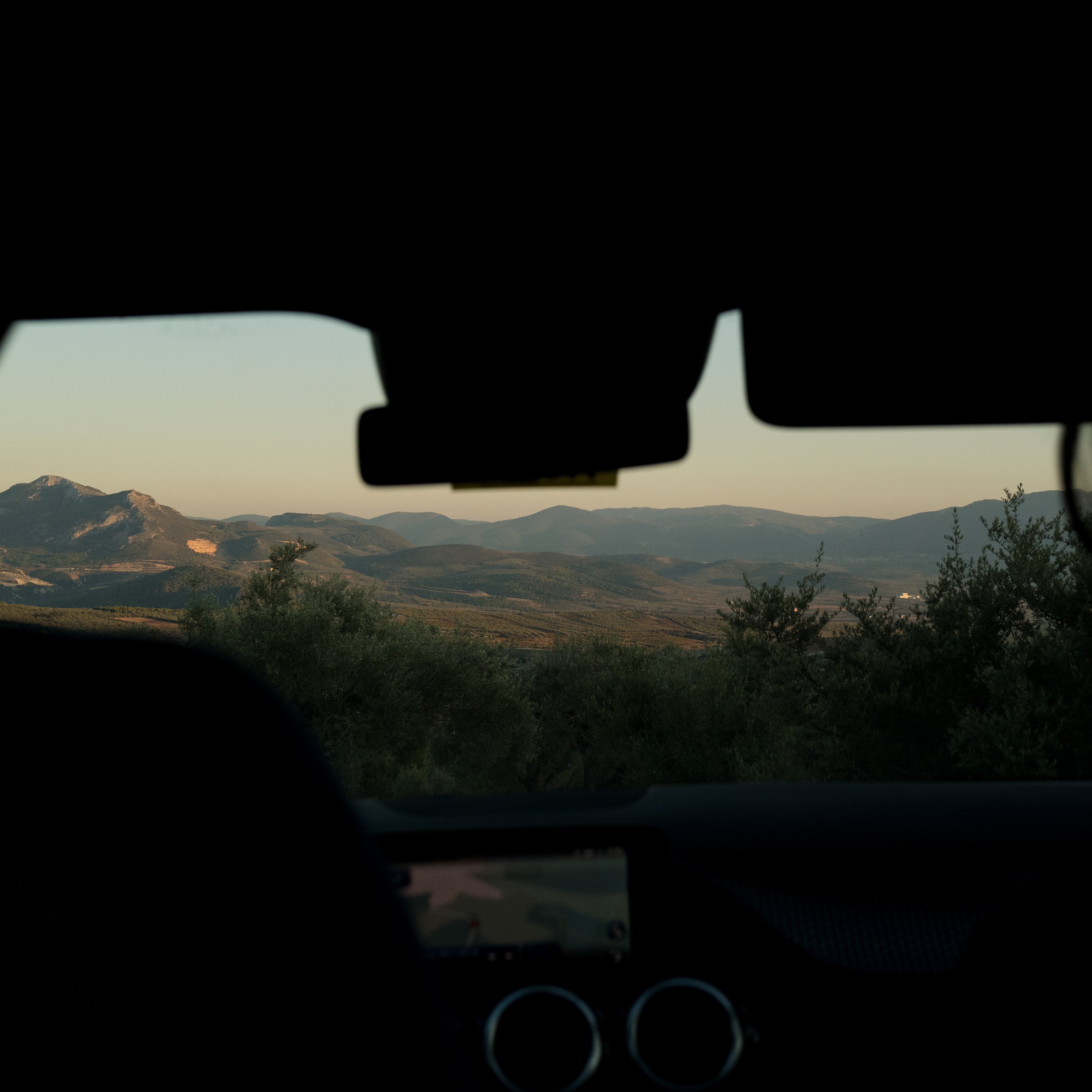 A view of rolling hills and mountains at dusk, seen from the silhouetted interior of a car.