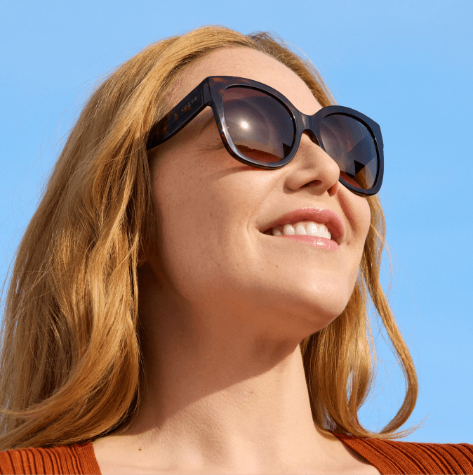 Une femme souriante portant de grandes lunettes de soleil écaille de tortue regarde vers le haut sous un ciel bleu dégagé.