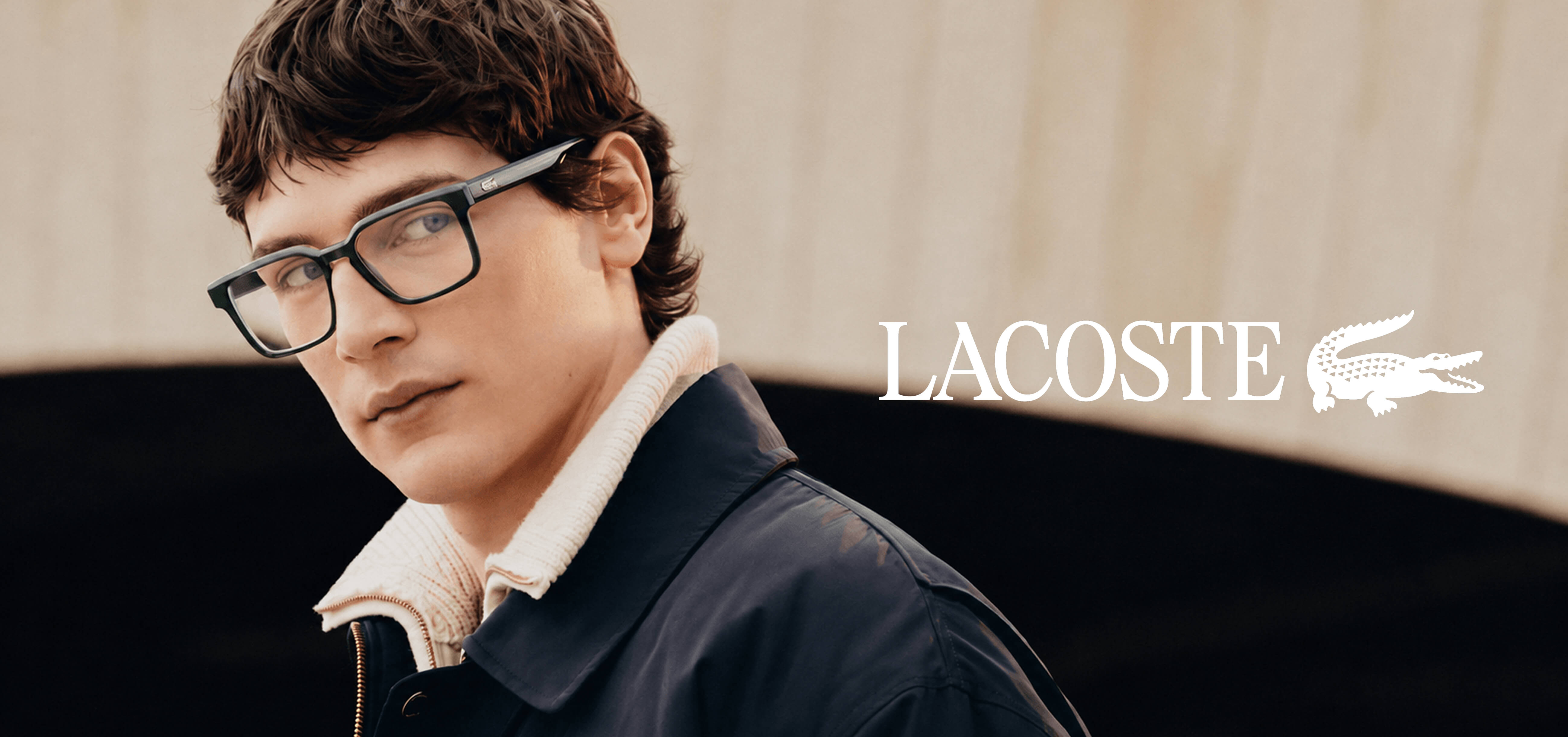 Un jeune homme aux cheveux foncés et aux yeux bleus clairs porte une paire de lunettes rectangulaires vert foncé Lacoste.