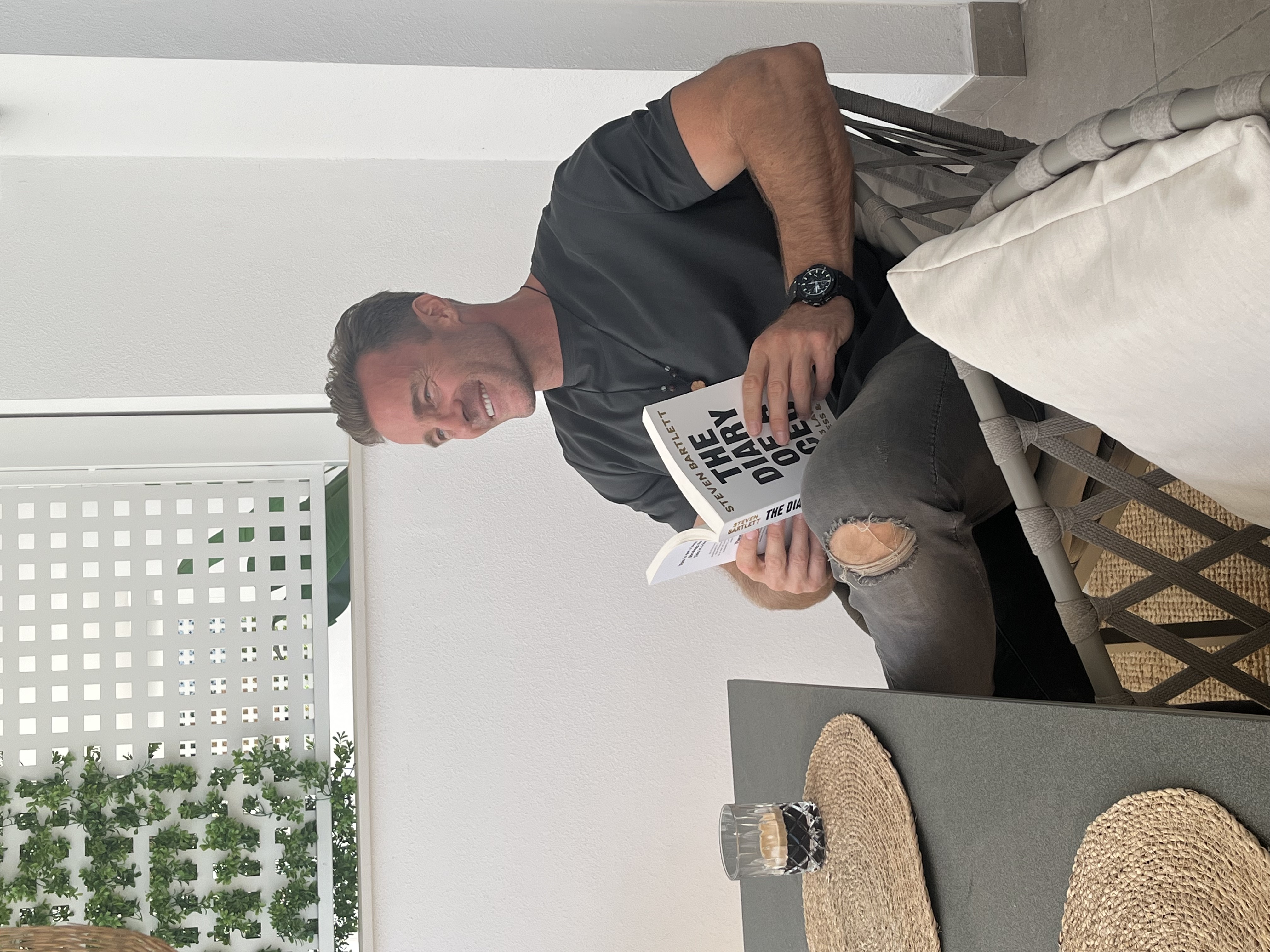 Person sitting outdoors, smiling, reading a book titled 'The Diary of a CEO', with a drink nearby.