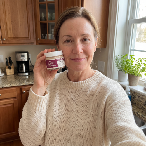 A woman in a cream-colored sweater holds up a small jar while taking a selfie in a kitchen.