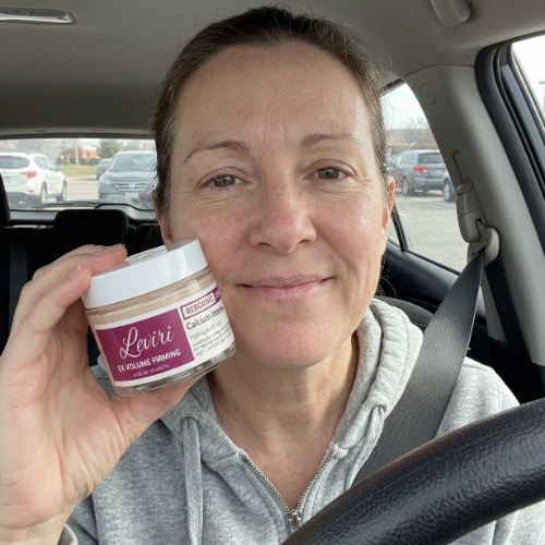 A woman in a car holds a jar of Leviri face cream next to her cheek and smiles.