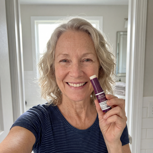 A smiling woman in a bathroom holds up a purple tube of a skincare product.