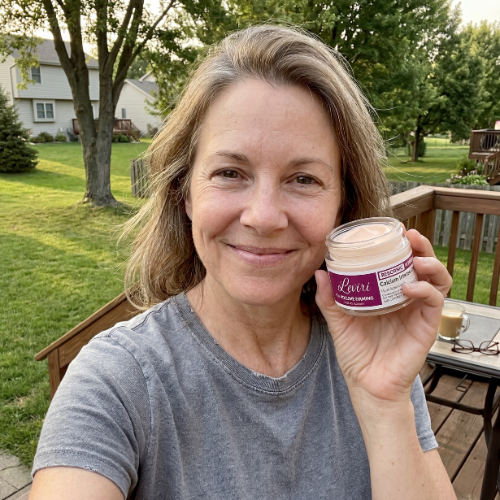 A smiling woman sits on a deck and holds up a small jar of cream.