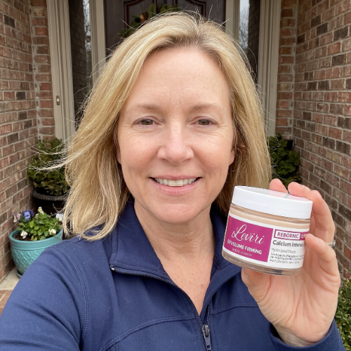 A smiling woman in a blue jacket stands outside holding a small jar of face cream.