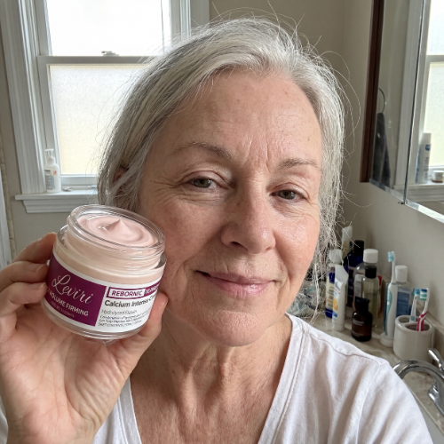 A woman with gray hair holds an open jar of pink face cream next to her cheek.