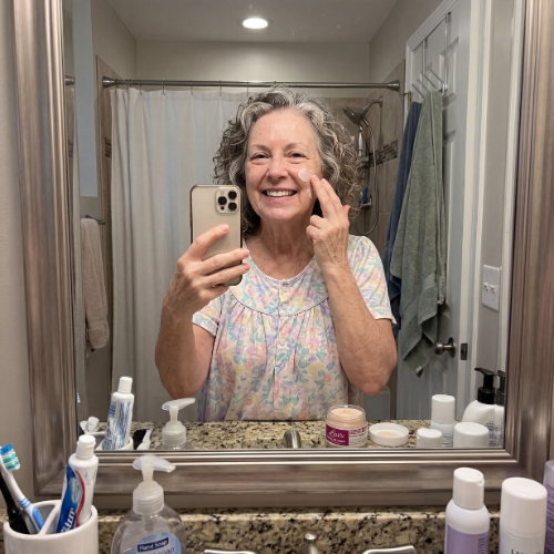 Woman taking a selfie in a bathroom mirror, applying cream to her face.