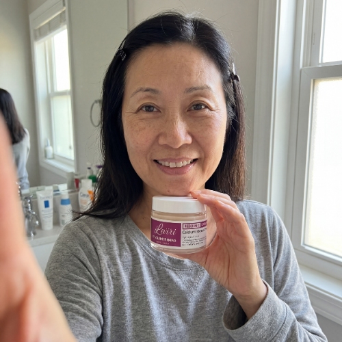 A smiling woman in a grey shirt holds up a small jar of face cream in a bathroom.