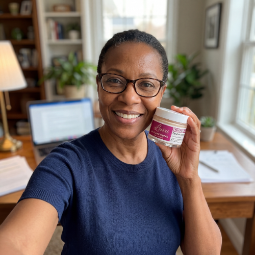 A smiling woman wearing glasses holds up a small jar of a product while sitting at her desk.