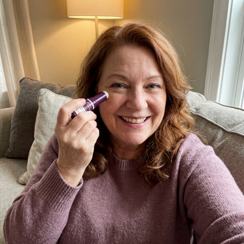 A smiling woman with red hair applies a purple eye treatment stick to the skin under her eye.