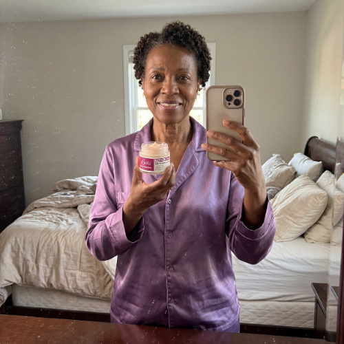A woman in purple satin pajamas takes a mirror selfie while holding a small jar of product.