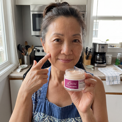A woman in a kitchen points to her cheek while holding a small jar of pink cream.