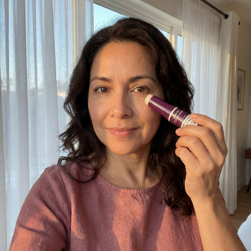 A woman with wavy dark hair applies a cosmetic product from a purple tube under her eye.