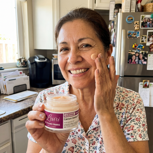 A smiling woman in a kitchen holds a jar of cream while applying it to her face.
