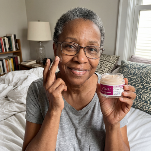 A smiling woman with glasses applies face cream to her cheek while sitting on a bed.