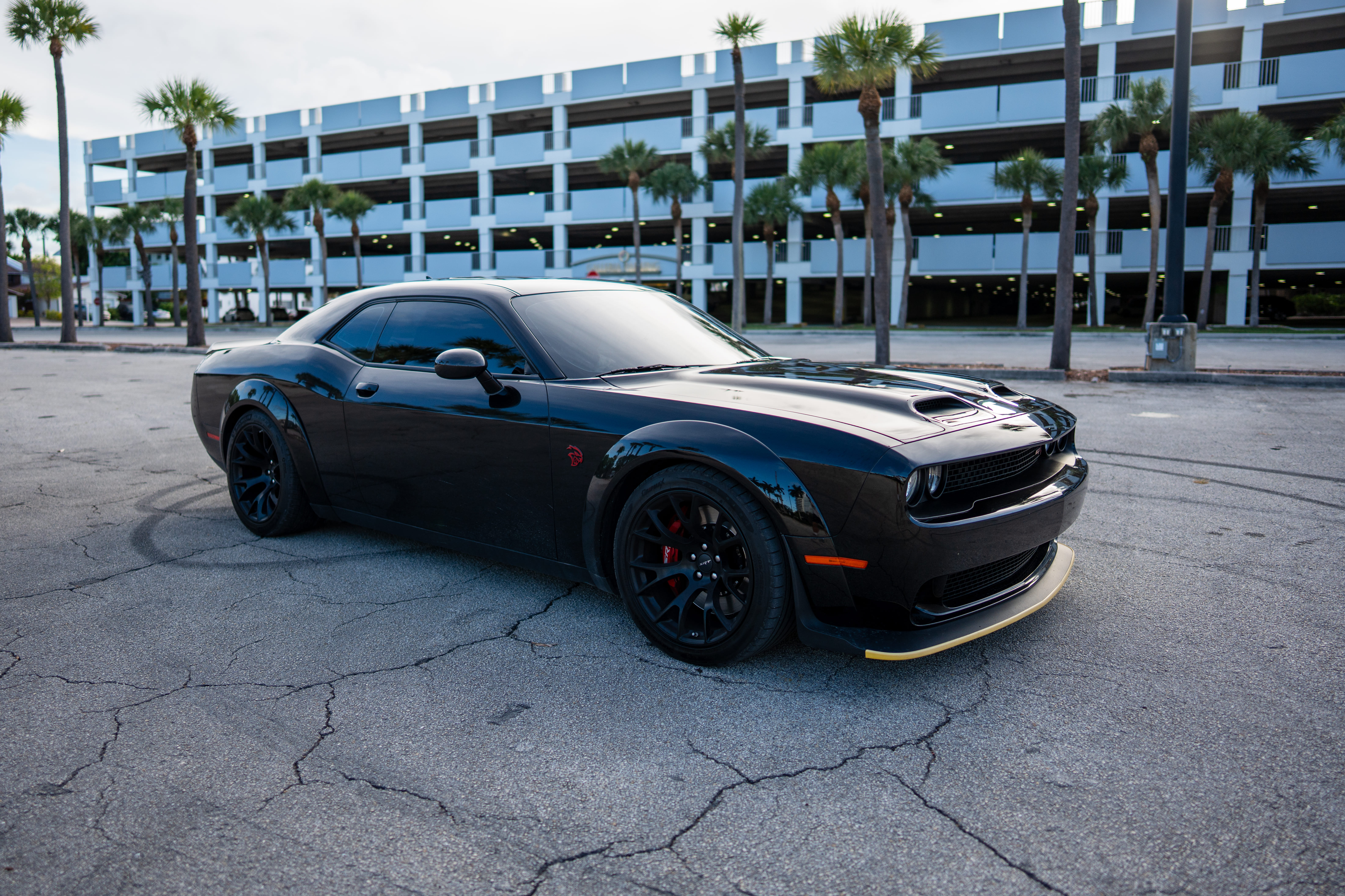 A black Dodge Challenger parked in a cracked asphalt lot with a parking garage and palm trees behind.