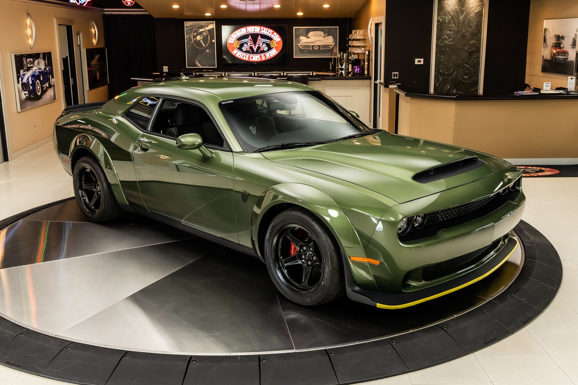 A green Dodge Challenger on a rotating display platform inside a car showroom.
