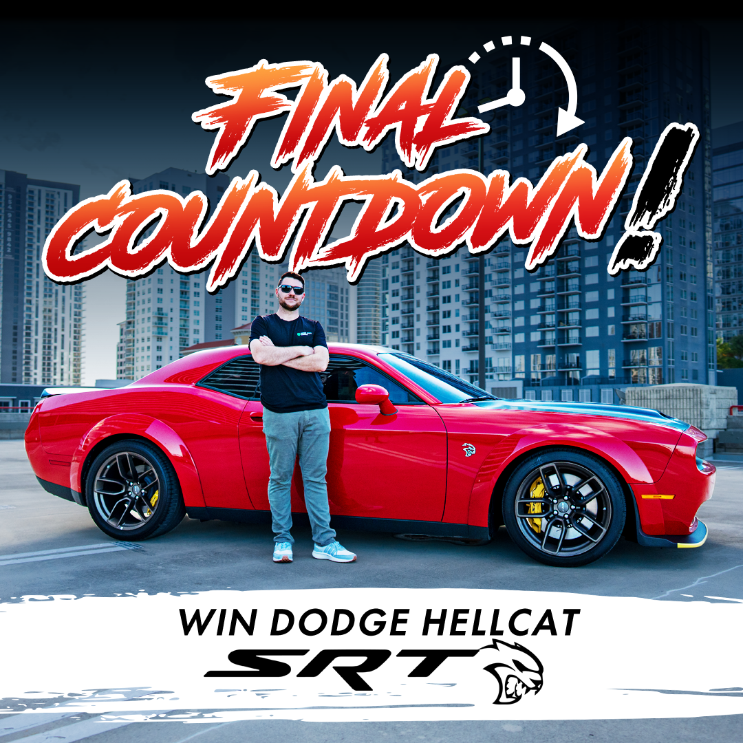 A man stands with his arms crossed next to a red Dodge Hellcat SRT in a city setting.
