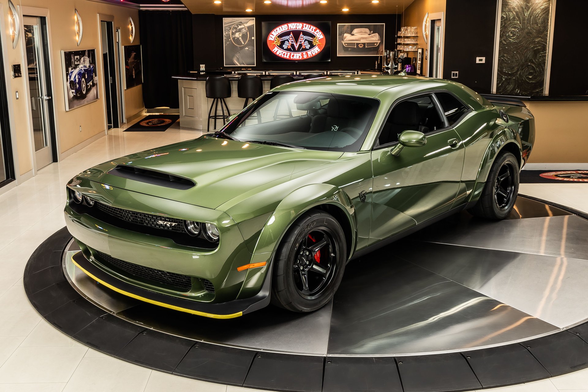 An olive green Dodge Challenger SRT on a rotating display platform inside a showroom.