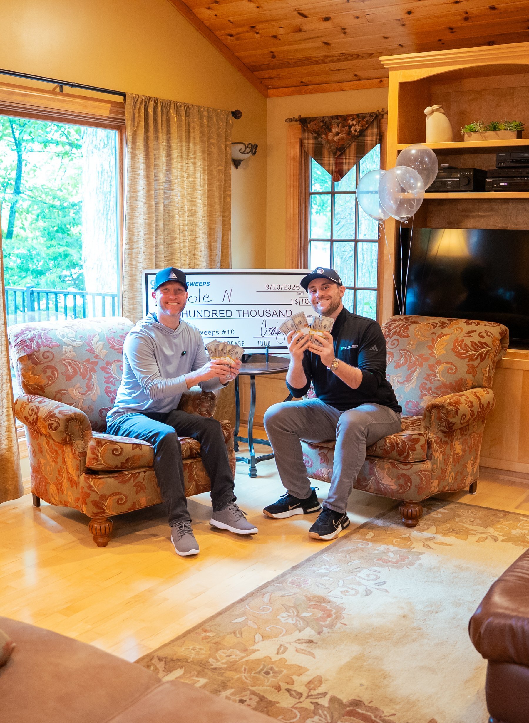 Two men sit in armchairs holding stacks of cash with a large novelty check between them.